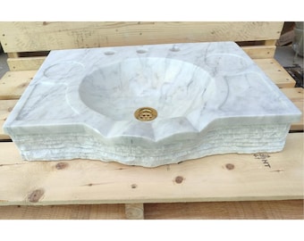 Pode incluir: Uma pia de banheiro de mármore branco com uma bacia arredondada e uma borda decorativa. A pia tem um ralo dourado e três orifícios para torneiras. A pia é feita de mármore branco com veios cinzentos.