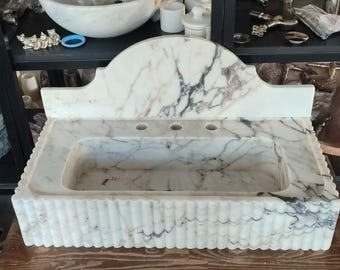 May include: A white marble sink with gray veining. The sink has a rectangular basin with a scalloped front edge and a curved backsplash. Three faucet holes are visible. A matching marble bowl sits on a shelf above.