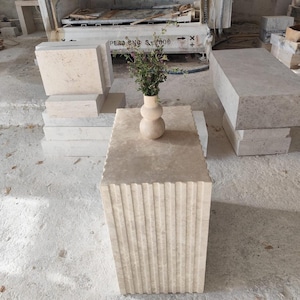 Puede incluir: Una mesa de centro de piedra beige con lados ranurados verticales. Un pequeño jarrón beige de tres secciones contiene un pequeño ramo de vegetación. Otros bloques de piedra y un largo banco de piedra están en el fondo.