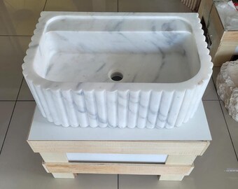 Pode incluir: Uma pia retangular de mármore branco com um design exterior canelado. A pia tem um orifício de drenagem central e está colocada sobre uma superfície branca em cima de uma caixa de madeira. O mármore apresenta veios cinzentos subtis.