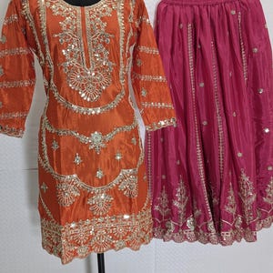 Puede incluir: Un conjunto festivo compuesto por una t&uacute;nica de seda naranja con intrincados bordados dorados y adornos de espejo, combinada con una falda fucsia. La t&uacute;nica presenta un cuello redondo y mangas tres cuartos. La falda est&aacute; plisada y tambi&eacute;n adornada con bordados dorados. Un collar dorado est&aacute; en el maniqu&iacute;.