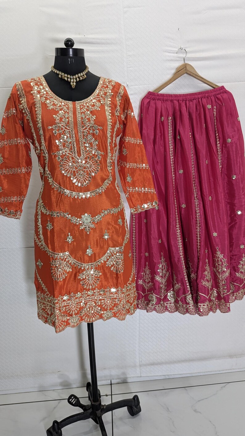 Puede incluir: Un conjunto festivo compuesto por una t&uacute;nica de seda naranja con intrincados bordados dorados y adornos de espejo, combinada con una falda fucsia. La t&uacute;nica presenta un cuello redondo y mangas tres cuartos. La falda est&aacute; plisada y tambi&eacute;n adornada con bordados dorados. Un collar dorado est&aacute; en el maniqu&iacute;.