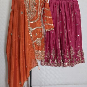 Puede incluir: Un conjunto de seda naranja con bordados y adornos dorados. El conjunto incluye una blusa de manga larga con un chal drapeado y una falda fucsia. El atuendo se exhibe en un maniqu&iacute; y una percha.
