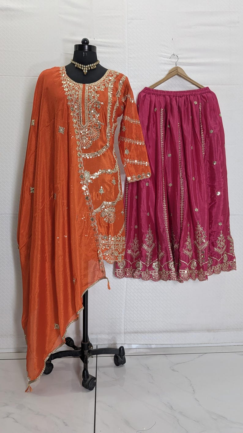 Puede incluir: Un conjunto de seda naranja con bordados y adornos dorados. El conjunto incluye una blusa de manga larga con un chal drapeado y una falda fucsia. El atuendo se exhibe en un maniqu&iacute; y una percha.