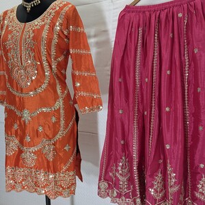 Puede incluir: Un conjunto de t&uacute;nica naranja y falda rosa. La t&uacute;nica presenta intrincados bordados y adornos dorados, con una falda a juego colgada de una percha de madera. El atuendo est&aacute; dise&ntilde;ado con una est&eacute;tica tradicional, adecuado para ocasiones festivas.