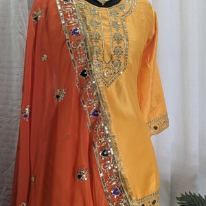May include: A bright yellow long-sleeved tunic with gold embroidery around the neckline and cuffs, paired with a vibrant orange dupatta featuring intricate gold floral patterns, sequin embellishments, and dark purple heart-shaped accents.