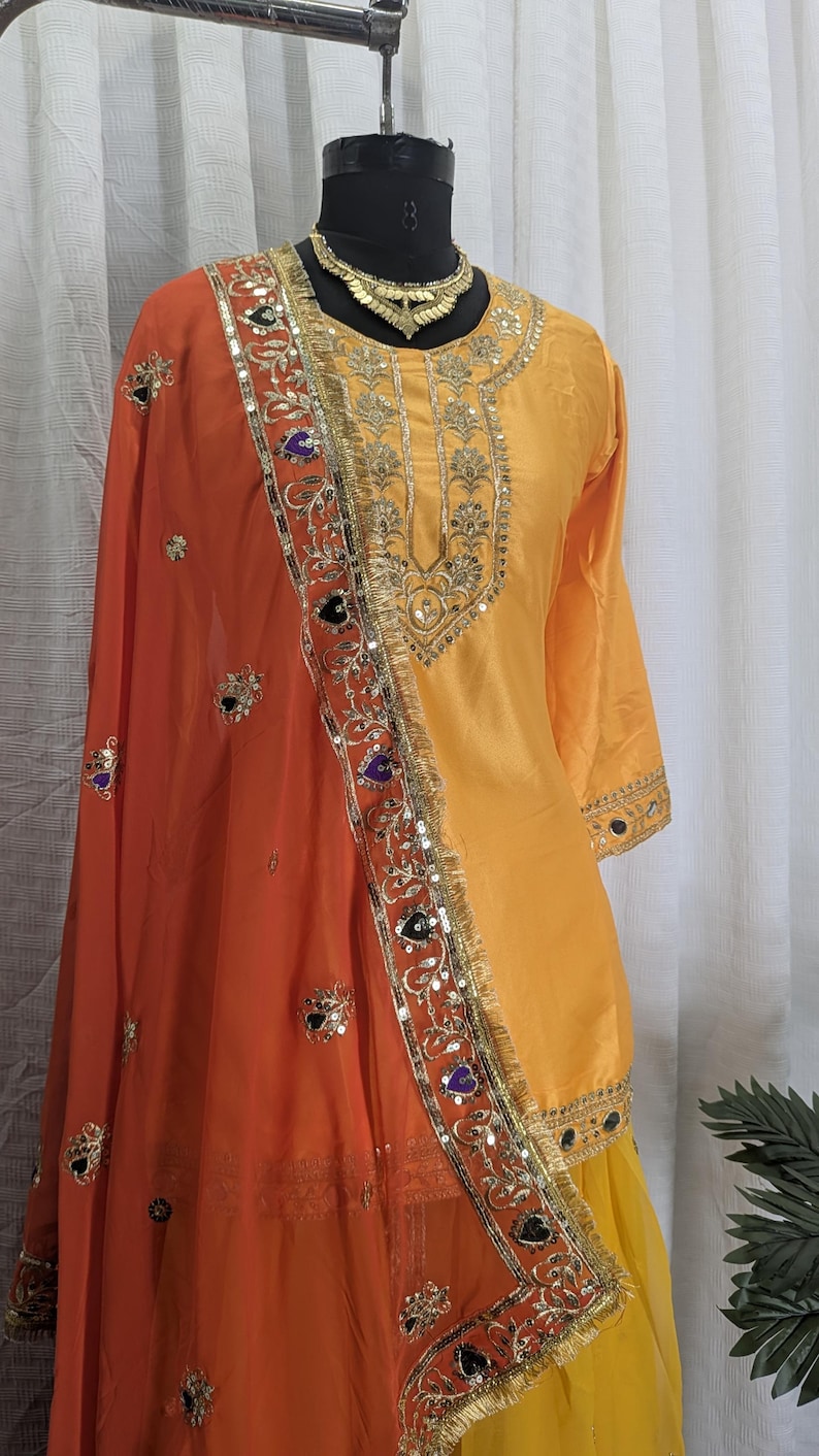 May include: A bright yellow long-sleeved tunic with gold embroidery around the neckline and cuffs, paired with a vibrant orange dupatta featuring intricate gold floral patterns, sequin embellishments, and dark purple heart-shaped accents.