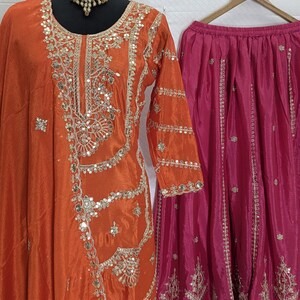 Puede incluir: Una t&uacute;nica de seda naranja con intrincados bordados y adornos plateados, combinada con una falda fucsia. La t&uacute;nica se exhibe en un maniqu&iacute; y la falda cuelga de una percha de madera. Tambi&eacute;n se incluye un collar a juego.