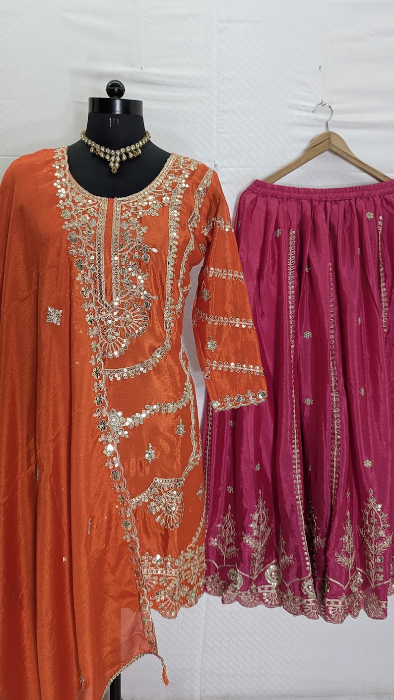 Puede incluir: Una t&uacute;nica de seda naranja con intrincados bordados y adornos plateados, combinada con una falda fucsia. La t&uacute;nica se exhibe en un maniqu&iacute; y la falda cuelga de una percha de madera. Tambi&eacute;n se incluye un collar a juego.