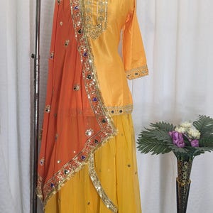 May include: A yellow three-piece outfit consisting of a long-sleeved tunic, a long skirt, and an orange dupatta. The tunic and skirt feature gold embroidery and mirror work. The orange dupatta has a wide gold border with colorful mirror accents.