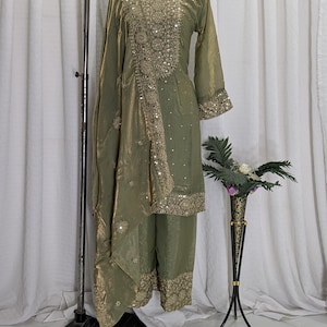 Peut inclure: Ensemble de vêtements de cérémonie vert olive. La tenue comprend une tunique à manches longues avec une broderie dorée complexe, un pantalon assorti et une écharpe fluide. La tenue est présentée sur un mannequin avec un collier noir. Un vase décoratif avec des fleurs est à droite.