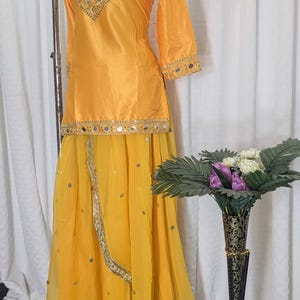May include: A bright yellow two-piece outfit consisting of a long-sleeved tunic and a long skirt. The tunic features gold embroidery and mirror work around the neckline and hem. The skirt has a sheer overlay with scattered mirror accents.