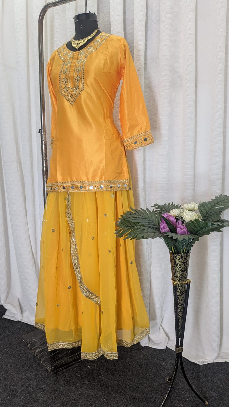 May include: A bright yellow two-piece outfit consisting of a long-sleeved tunic and a long skirt. The tunic features gold embroidery and mirror work around the neckline and hem. The skirt has a sheer overlay with scattered mirror accents.