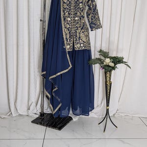 May include: Navy blue embroidered Indian suit with gold accents. The ensemble includes a long-sleeved top, wide-leg pants, and a matching scarf. A gold necklace is displayed on the mannequin. A decorative black and gold vase with flowers is on the right.
