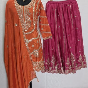 Puede incluir: Un conjunto de ropa que incluye una t&uacute;nica de seda naranja con bordados dorados y lentejuelas, y una falda fucsia. La t&uacute;nica tiene mangas largas y un dise&ntilde;o drapeado. La falda est&aacute; adornada con bordados dorados. Un collar dorado est&aacute; en el maniqu&iacute;.