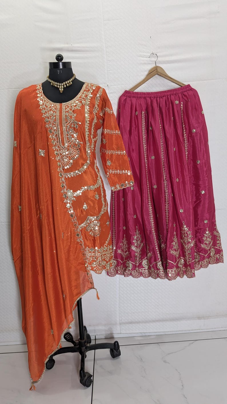 Puede incluir: Un conjunto de ropa que incluye una t&uacute;nica de seda naranja con bordados dorados y lentejuelas, y una falda fucsia. La t&uacute;nica tiene mangas largas y un dise&ntilde;o drapeado. La falda est&aacute; adornada con bordados dorados. Un collar dorado est&aacute; en el maniqu&iacute;.