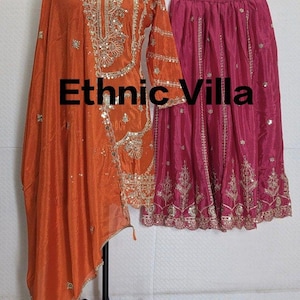 Puede incluir: Un conjunto de t&uacute;nica y dupatta de seda naranja con intrincados bordados y adornos dorados. Una falda rosa fucsia con bordados a juego cuelga cerca. El texto "Ethnic Villa" se muestra en la imagen.