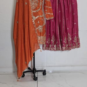 Puede incluir: Un conjunto &eacute;tnico naranja y fucsia. La parte superior naranja presenta intrincados bordados dorados y adornos de lentejuelas. La falda rosa fucsia tambi&eacute;n est&aacute; adornada con bordados dorados. El conjunto se exhibe en un maniqu&iacute; y una percha.