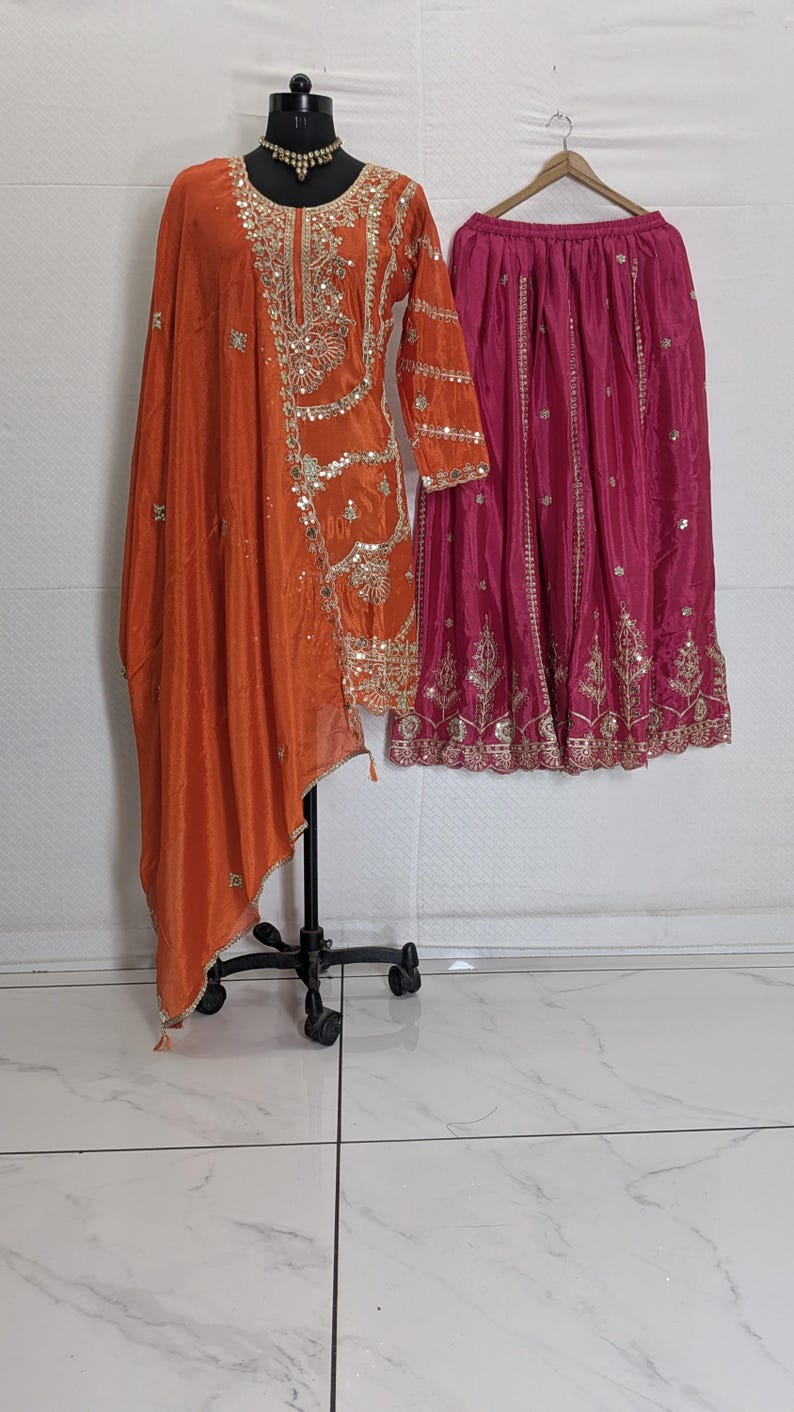 Puede incluir: Un conjunto &eacute;tnico naranja y fucsia. La parte superior naranja presenta intrincados bordados dorados y adornos de lentejuelas. La falda rosa fucsia tambi&eacute;n est&aacute; adornada con bordados dorados. El conjunto se exhibe en un maniqu&iacute; y una percha.