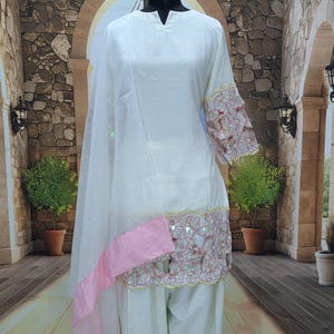 May include: A white three-piece outfit consisting of a long-sleeved tunic, loose-fitting trousers, and a matching dupatta. The tunic features a V-neckline and intricate floral cutwork embroidery on the sleeves and hem, accented with pink fabric trim.