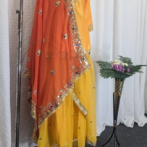 May include: A bright yellow long-sleeved tunic and matching skirt set paired with a vibrant orange dupatta. The ensemble features intricate gold embroidery, metallic trim, and small reflective mirror accents along the borders and neckline.