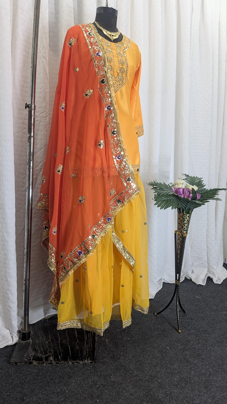 May include: A bright yellow long-sleeved tunic and matching skirt set paired with a vibrant orange dupatta. The ensemble features intricate gold embroidery, metallic trim, and small reflective mirror accents along the borders and neckline.