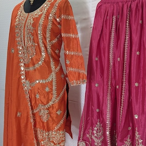 Puede incluir: Un conjunto de ropa tradicional del sur de Asia, que incluye una t&uacute;nica de seda naranja con intrincados bordados dorados y espejos, y una falda rosa fucsia con bordados dorados similares. Las prendas se exhiben en maniqu&iacute;es.