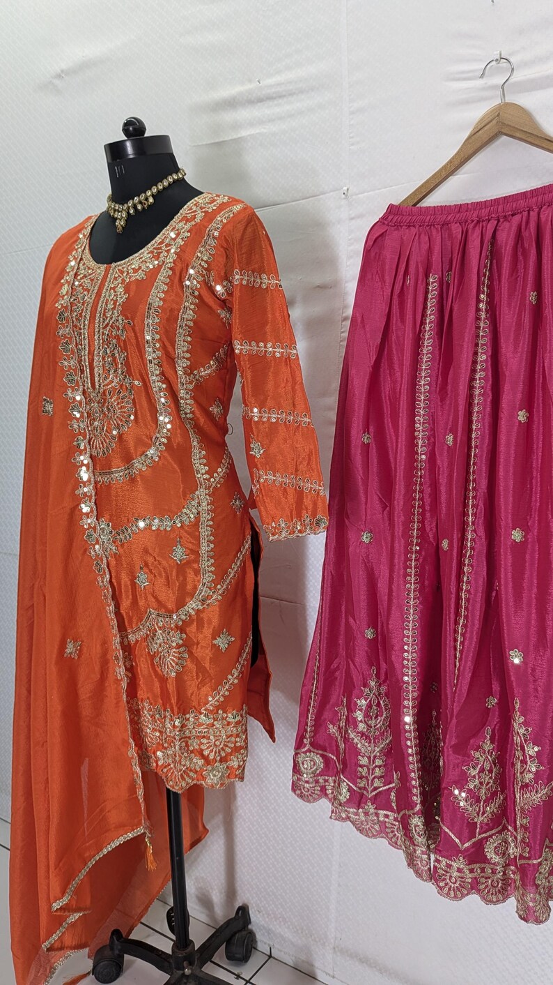 Puede incluir: Un conjunto de ropa tradicional del sur de Asia, que incluye una t&uacute;nica de seda naranja con intrincados bordados dorados y espejos, y una falda rosa fucsia con bordados dorados similares. Las prendas se exhiben en maniqu&iacute;es.