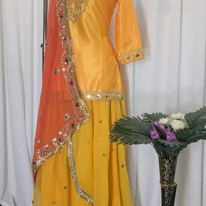 May include: A bright yellow three-piece outfit featuring a long-sleeved tunic with gold embroidery and mirror work, a matching flowing skirt, and a sheer orange dupatta with an ornate gold border, sequins, and colorful mirror embellishments.