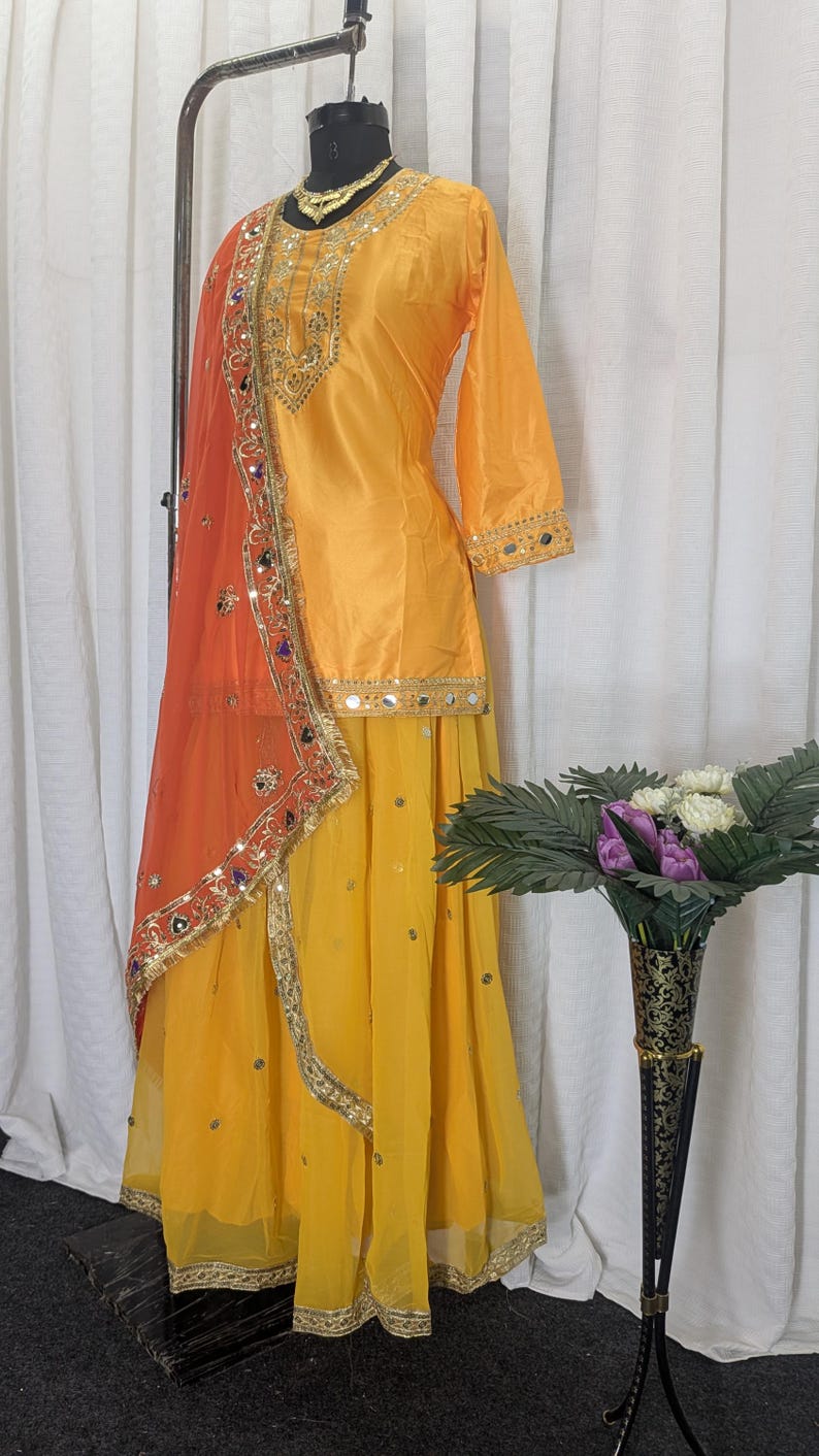 May include: A bright yellow three-piece outfit featuring a long-sleeved tunic with gold embroidery and mirror work, a matching flowing skirt, and a sheer orange dupatta with an ornate gold border, sequins, and colorful mirror embellishments.