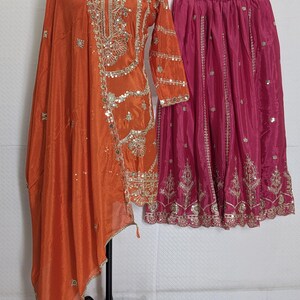 Puede incluir: Un conjunto de t&uacute;nica y dupatta de seda naranja con intrincados bordados y adornos dorados. Una falda rosa fucsia con bordados dorados a juego cuelga cerca. El atuendo se exhibe en un maniqu&iacute;, mostrando el dise&ntilde;o tradicional indio.