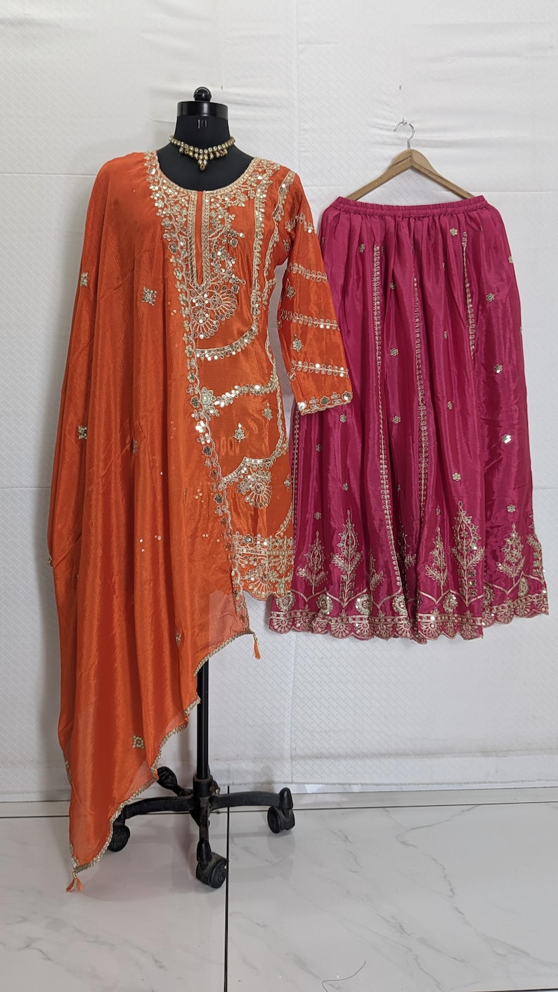 Puede incluir: Un conjunto de t&uacute;nica y dupatta de seda naranja con intrincados bordados y adornos dorados. Una falda rosa fucsia con bordados dorados a juego cuelga cerca. El atuendo se exhibe en un maniqu&iacute;, mostrando el dise&ntilde;o tradicional indio.