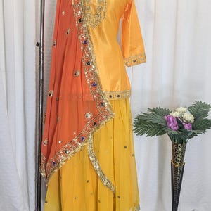 May include: A yellow three-piece outfit consisting of a long-sleeved tunic, a flowing skirt, and an orange dupatta. The tunic and skirt feature gold embroidery and small mirrors. The orange dupatta has a wide gold border with colorful accents.