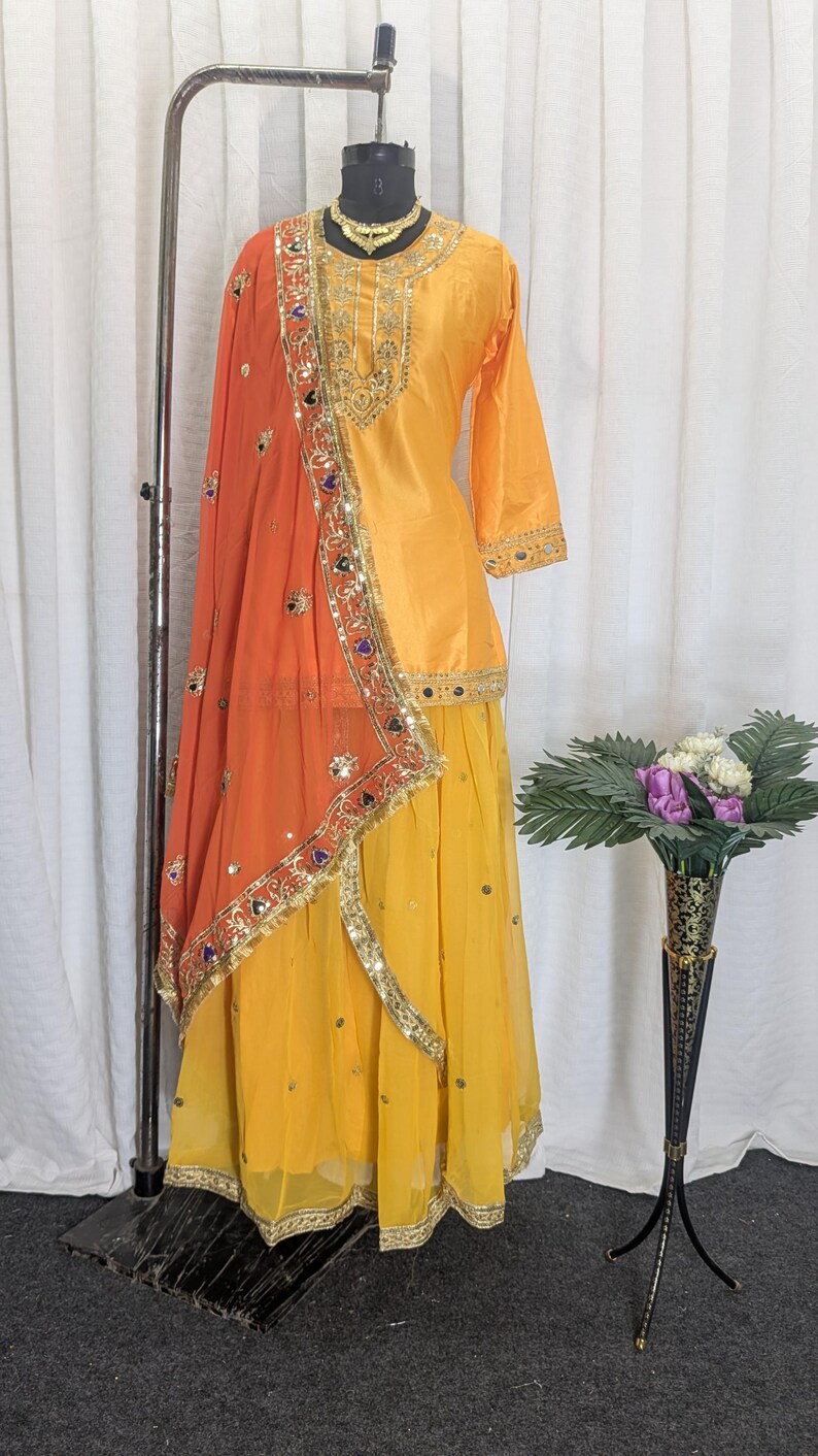 May include: A yellow three-piece outfit consisting of a long-sleeved tunic, a flowing skirt, and an orange dupatta. The tunic and skirt feature gold embroidery and small mirrors. The orange dupatta has a wide gold border with colorful accents.