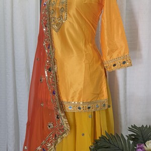 May include: A bright yellow tunic with three-quarter sleeves and a matching yellow skirt. The tunic features gold embroidery and mirror work along the neckline and hem. An orange sheer dupatta with gold trim and colorful mirror accents is draped over.