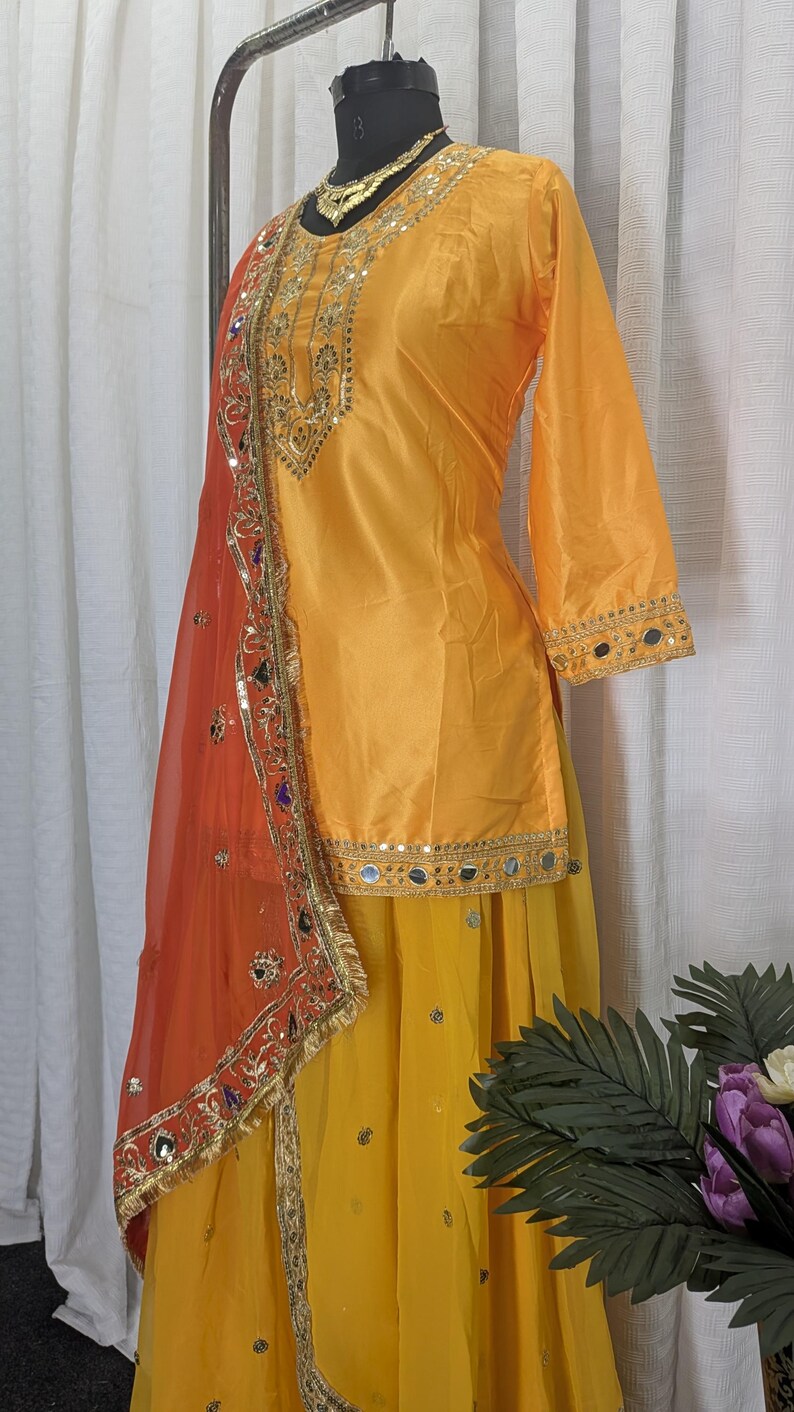 May include: A bright yellow tunic with three-quarter sleeves and a matching yellow skirt. The tunic features gold embroidery and mirror work along the neckline and hem. An orange sheer dupatta with gold trim and colorful mirror accents is draped over.