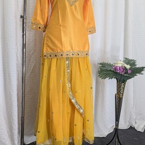 May include: A bright yellow two-piece outfit consisting of a long-sleeved tunic and a flowing skirt. The tunic features gold embroidery and mirror work around the neckline and cuffs. The skirt has a layered design with gold trim and mirror accents.