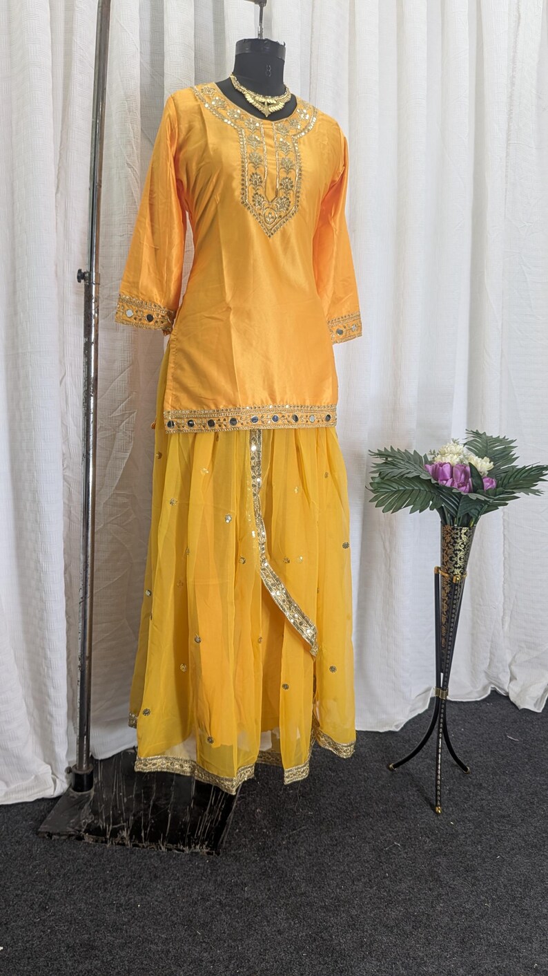 May include: A bright yellow two-piece outfit consisting of a long-sleeved tunic and a flowing skirt. The tunic features gold embroidery and mirror work around the neckline and cuffs. The skirt has a layered design with gold trim and mirror accents.