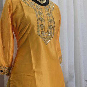 May include: A bright yellow tunic featuring three-quarter sleeves and a round neckline. The garment is adorned with intricate gold embroidery and reflective mirror embellishments along the neckline, sleeve cuffs, and the lower hemline.