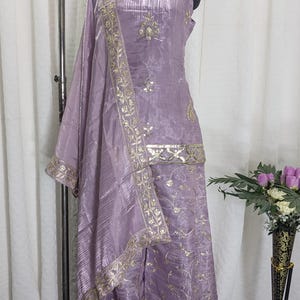 May include: A lavender-colored salwar kameez outfit displayed on a mannequin. The ensemble includes a sleeveless top, wide-leg pants, and a matching scarf. Gold embroidery and embellishments adorn the outfit, with a decorative border on the scarf and pants.