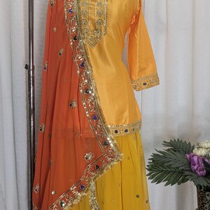 May include: A bright yellow three-piece outfit featuring a long-sleeved tunic with gold embroidery, a matching yellow skirt, and a sheer orange dupatta. The ensemble is decorated with gold trim, small mirrors, and colorful accents along the borders.
