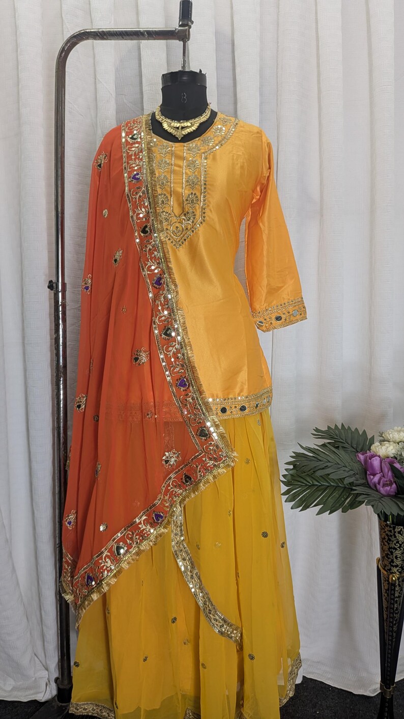 May include: A bright yellow three-piece outfit featuring a long-sleeved tunic with gold embroidery, a matching yellow skirt, and a sheer orange dupatta. The ensemble is decorated with gold trim, small mirrors, and colorful accents along the borders.