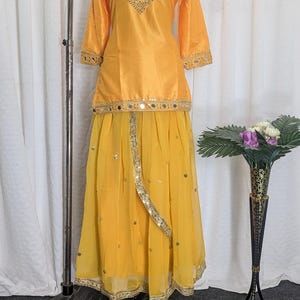 May include: A bright yellow two-piece outfit consisting of a long-sleeved tunic and a flowing skirt. The tunic features a round neckline with gold embroidery and mirror work. The skirt is made of sheer fabric with scattered mirror embellishments.