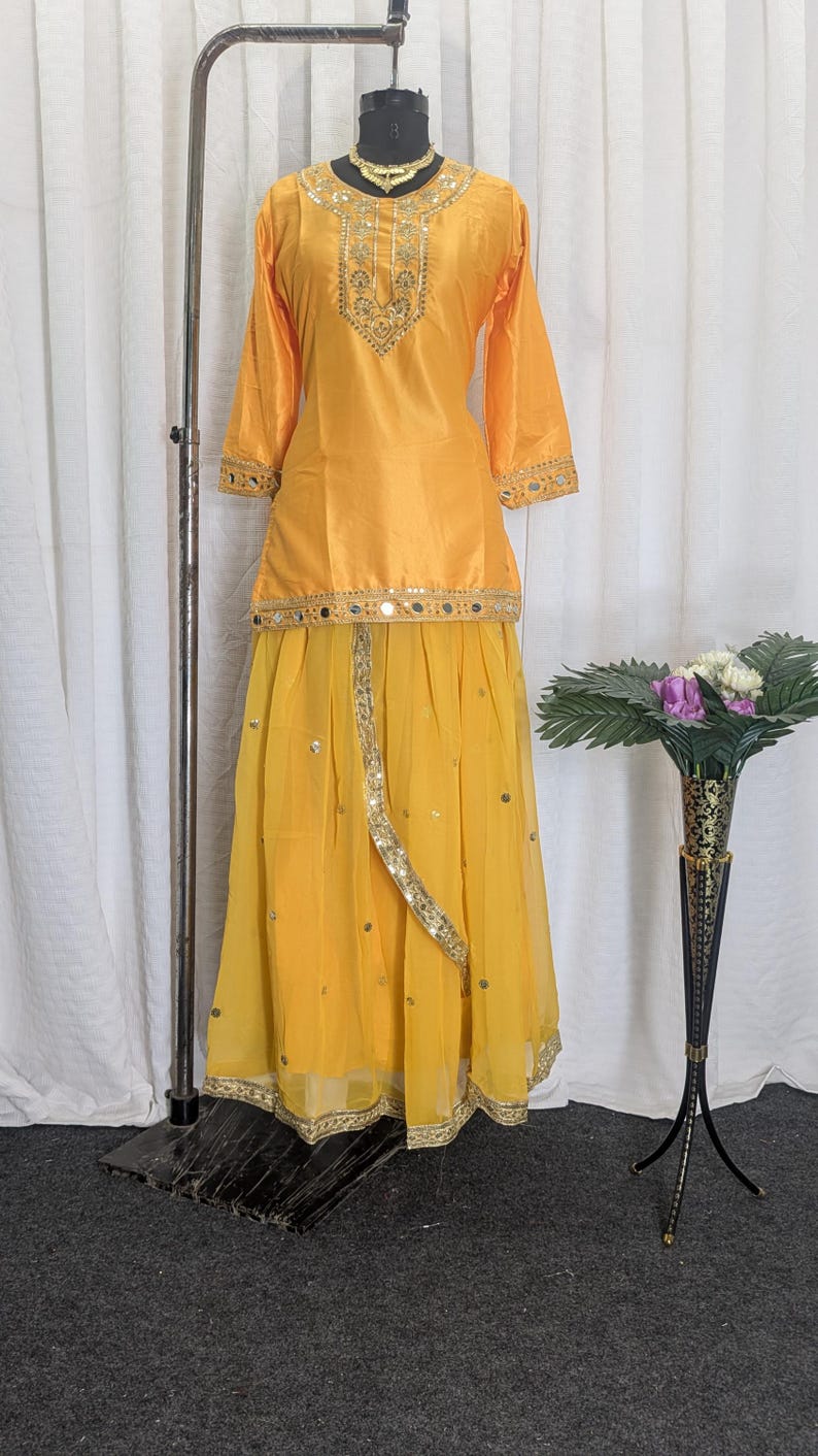 May include: A bright yellow two-piece outfit consisting of a long-sleeved tunic and a flowing skirt. The tunic features a round neckline with gold embroidery and mirror work. The skirt is made of sheer fabric with scattered mirror embellishments.