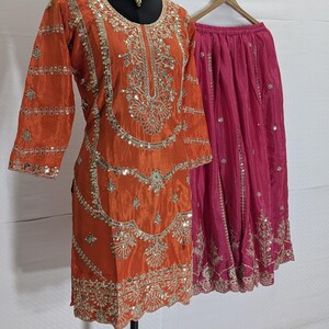 Puede incluir: Un conjunto de ropa, compuesto por una t&uacute;nica naranja y una falda rosa, se muestra en un maniqu&iacute; y una percha. La t&uacute;nica est&aacute; adornada con intrincados bordados dorados y lentejuelas. La falda es de color rosa vibrante con bordados a juego.