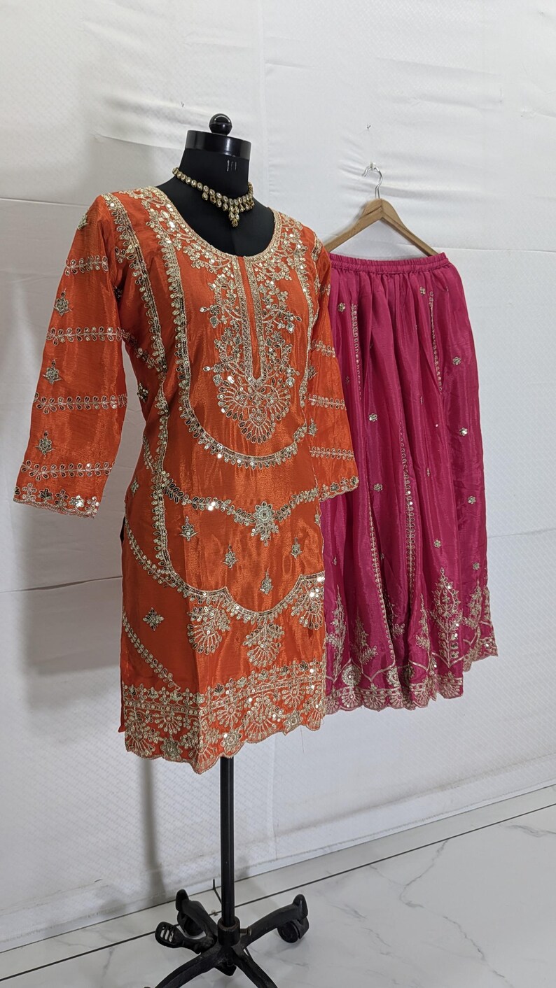 Puede incluir: Un conjunto de ropa, compuesto por una t&uacute;nica naranja y una falda rosa, se muestra en un maniqu&iacute; y una percha. La t&uacute;nica est&aacute; adornada con intrincados bordados dorados y lentejuelas. La falda es de color rosa vibrante con bordados a juego.
