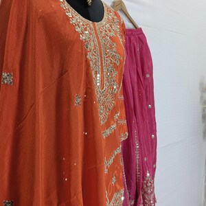 Puede incluir: Un conjunto de t&uacute;nica naranja bordada y falda rosa, exhibido en un maniqu&iacute;. La t&uacute;nica presenta intrincados bordados dorados y lentejuelas. Un collar dorado adorna el cuello del maniqu&iacute;. El atuendo es probablemente para una ocasi&oacute;n especial.