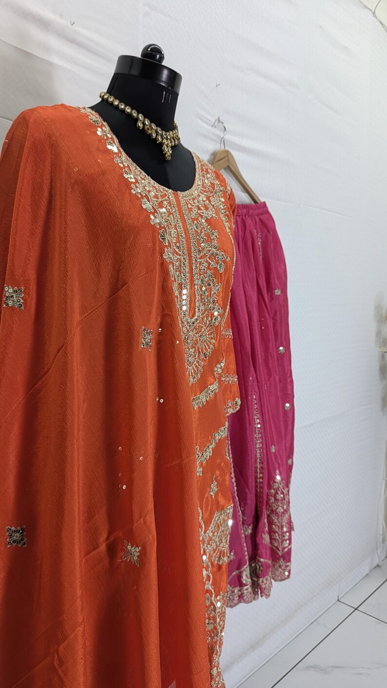 Puede incluir: Un conjunto de t&uacute;nica naranja bordada y falda rosa, exhibido en un maniqu&iacute;. La t&uacute;nica presenta intrincados bordados dorados y lentejuelas. Un collar dorado adorna el cuello del maniqu&iacute;. El atuendo es probablemente para una ocasi&oacute;n especial.