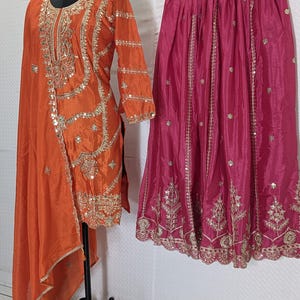 Puede incluir: Una t&uacute;nica de seda naranja con bordados y adornos dorados se exhibe en un maniqu&iacute;. Una falda fucsia con bordados dorados cuelga al lado. El conjunto probablemente forma parte de una colecci&oacute;n de ropa tradicional o formal.