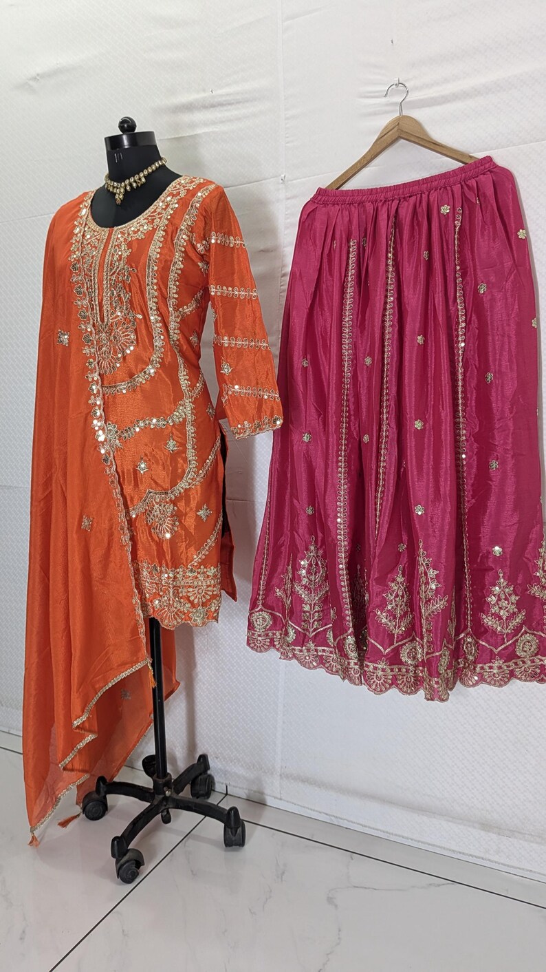 Puede incluir: Una t&uacute;nica de seda naranja con bordados y adornos dorados se exhibe en un maniqu&iacute;. Una falda fucsia con bordados dorados cuelga al lado. El conjunto probablemente forma parte de una colecci&oacute;n de ropa tradicional o formal.